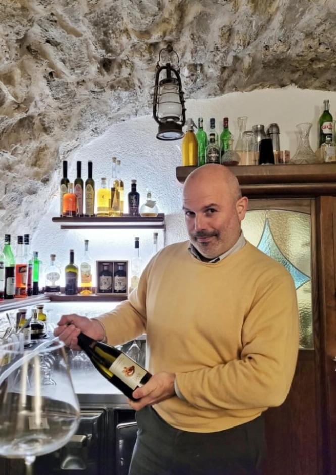Uomo sorridente in cantina rustica, bottiglia di vino in mano, atmosfera accogliente.